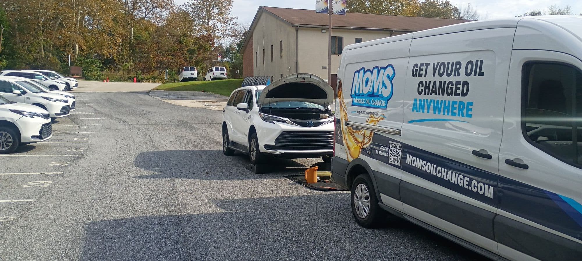 The MOMS Mobile Oil Change van beside a white Toyota Sienna with its hood up during a driveway oil change